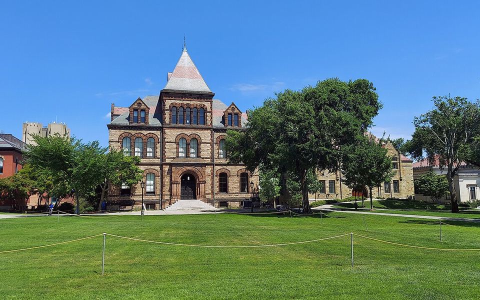 Sayles Hall, a stone hall with a pointed tower roof and arched windows, behind a wide lawn on Brown University&rsquo;s campus in Providence, Rhode Island