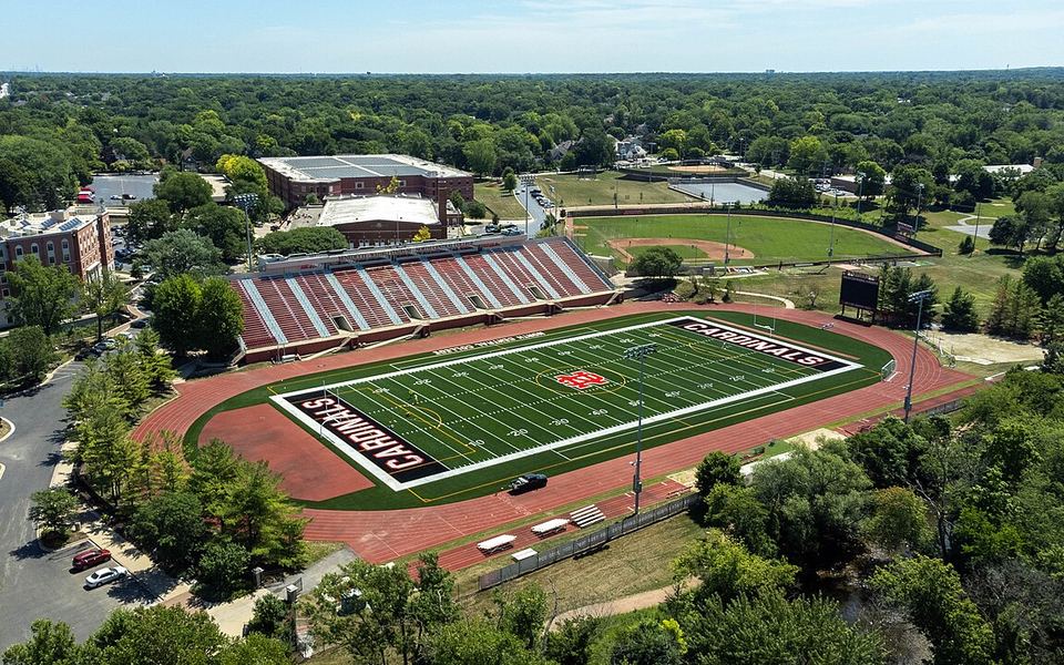 Aerial view of Benedetti-Wehrli Stadium on North Central College&rsquo;s campus in Naperville, Illinois: a football field with &ldquo;Cardinals&rdquo; end zones inside a red running track, with red-brick campus buildings, a baseball field and trees behind